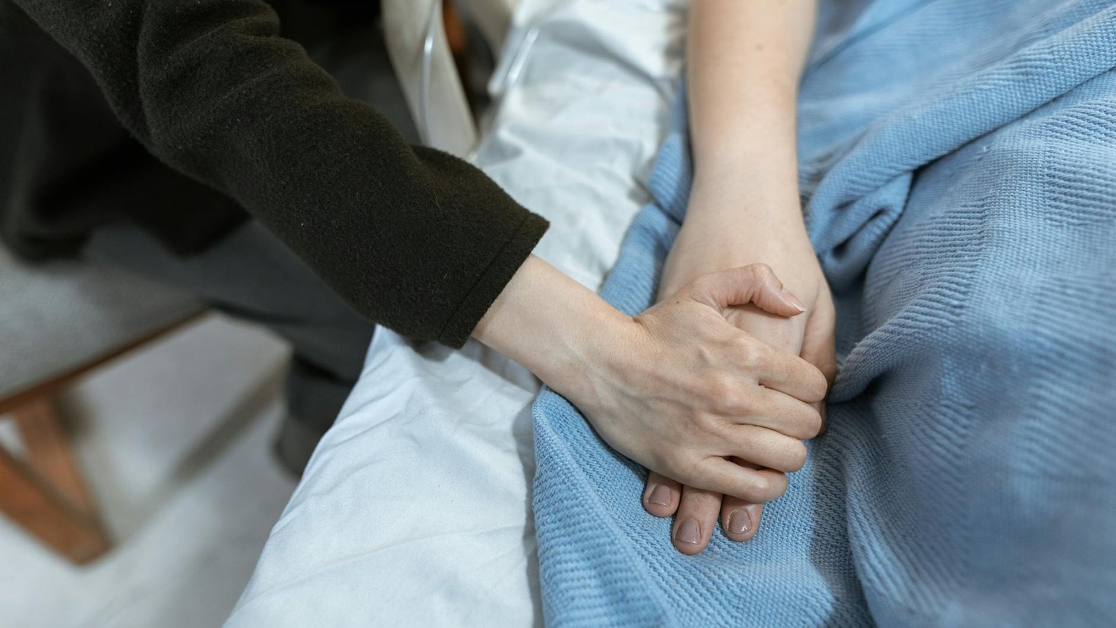 A caregiver's hand gently resting on a patient's clasped hands at a bedside