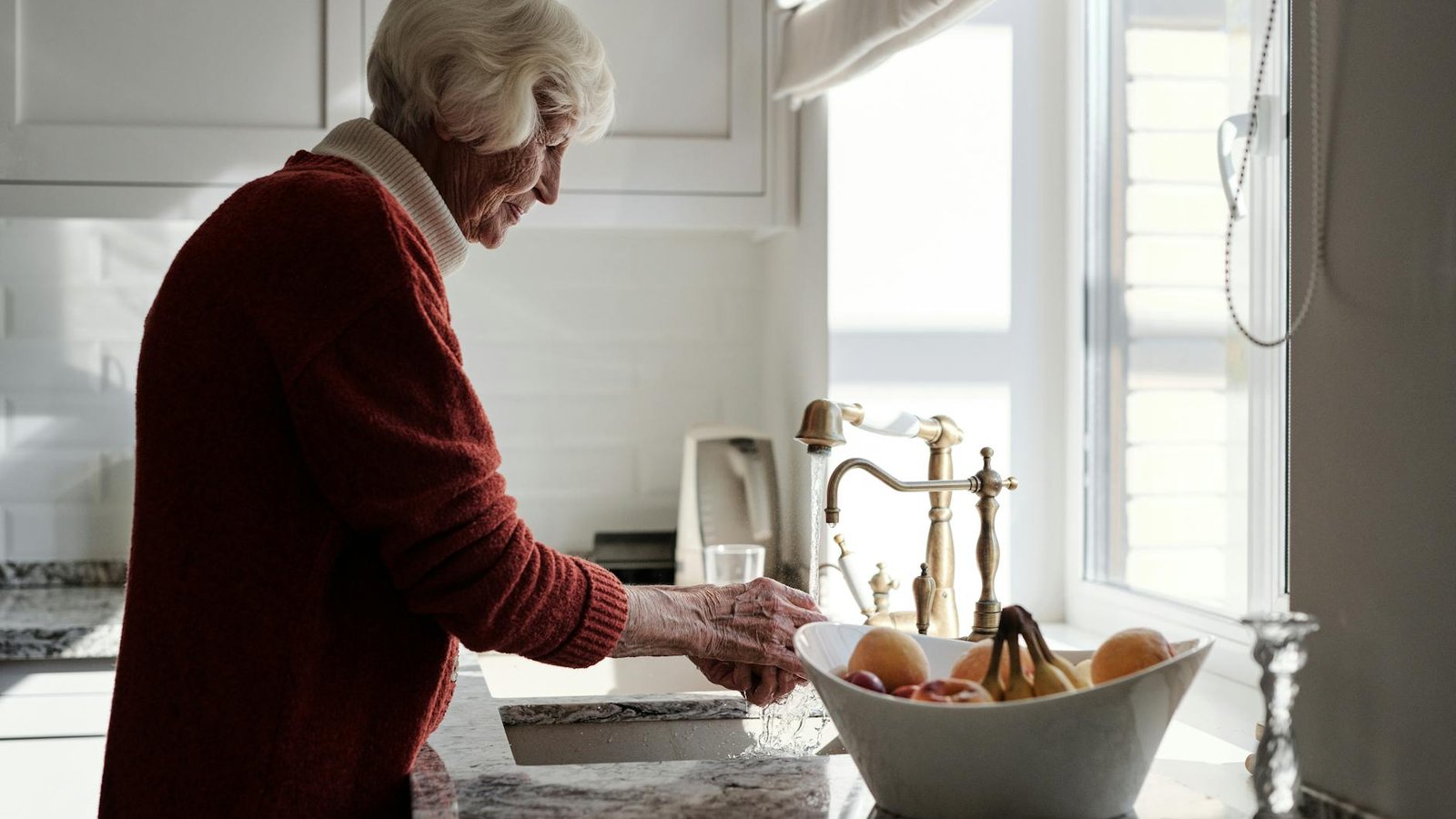 An older woman washing fruit at her kitchen sink in warm afternoon light